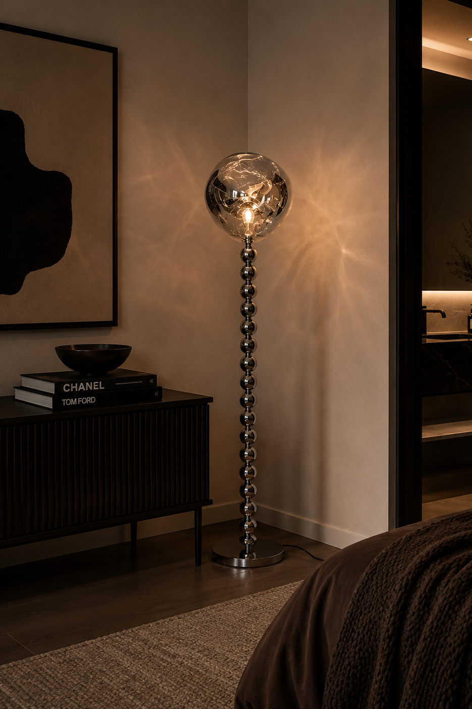 A modern floor lamp with a clear faceted glass globe shade and a metallic beaded stem, glowing in a warmly lit bedroom setting next to a dark console table.